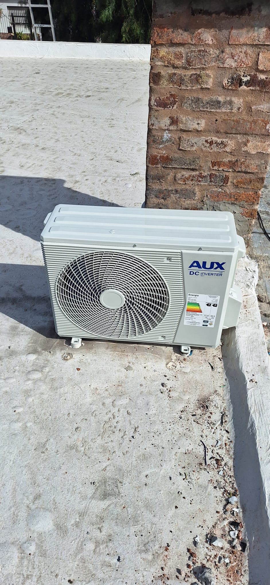 Unidad exterior AUX DC Inverter instalada en una azotea, apoyada sobre sus bases y alineada contra una pared de ladrillo.